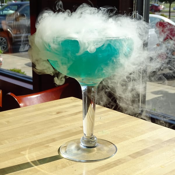 A large blue cocktail in a tall glass, emitting white smoke, sits on a light wooden table.