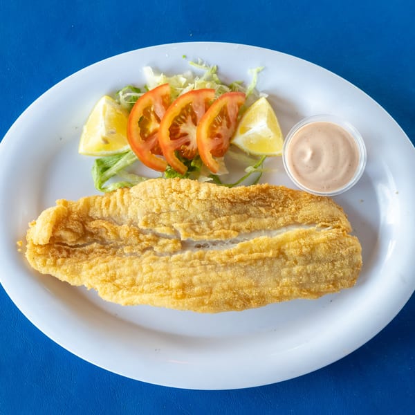Golden-fried fish fillet with a side of lettuce, tomato, lemon wedges, and a small cup of sauce.