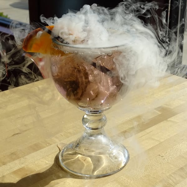 A smoking cocktail in a goblet, garnished with an orange slice, on a light wooden surface.