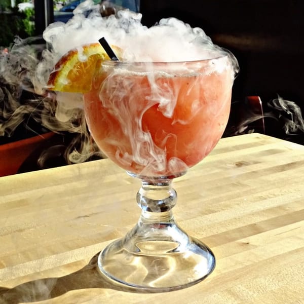 A smoking red cocktail with an orange slice and straw sits on a wooden table.
